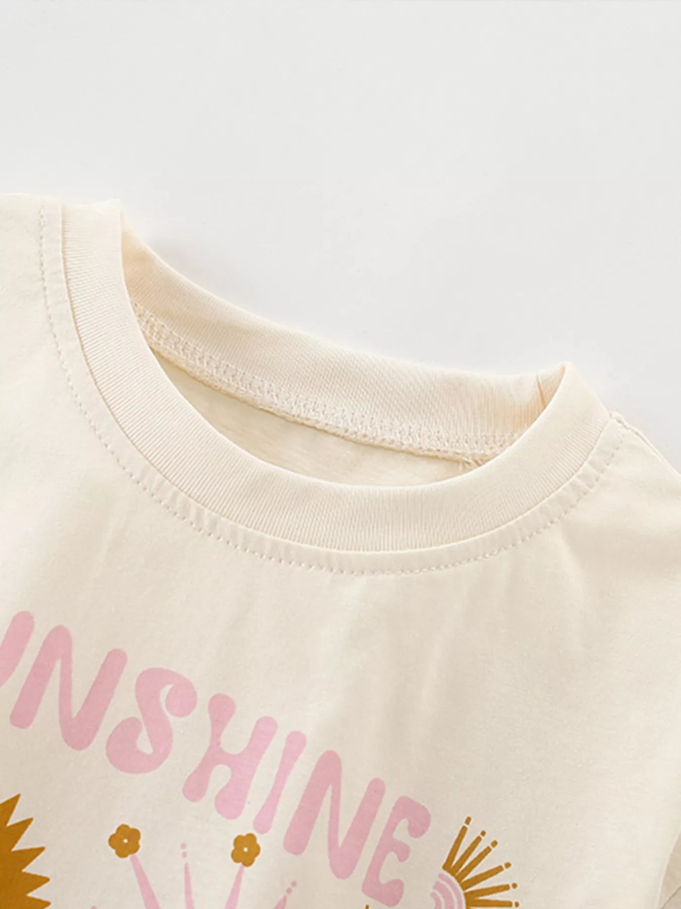 Beige t-shirt with pink 'SHINE' text and sunflower graphics on a light gray background